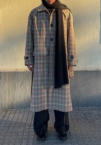 Kockás, hosszú kabát bézs, kék és piros színben, nagy gombokkal, gallérral és két oldalsó zsebbel, fekete bordázott sállal és nadrággal párosítva.