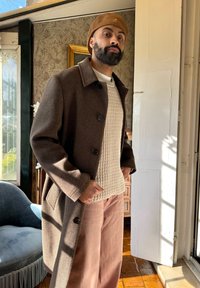 Rjav volnen plašč z velikimi gumbi, položen na bež teksturirano pulover. Usklajen s svetlo rožnatimi širokimi hlačami. Naravna svetloba poudarja teksture.