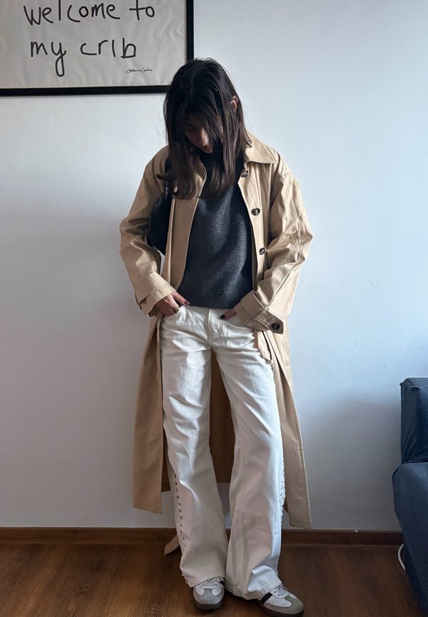 Persona che indossa un trench beige, maglione grigio scuro, pantaloni bianchi e scarpe da ginnastica, in piedi contro un muro bianco sotto un cartello con la scritta "benvenuti nella mia casa".