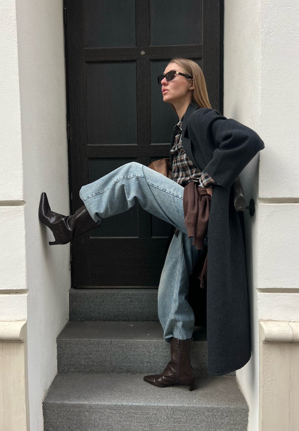 Femme portant des lunettes de soleil, un manteau sombre, une chemise à carreaux et un jean clair, assise sur des marches avec une jambe relevée, devant un mur blanc et une porte noire.