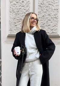 Jeune femme portant des lunettes et un manteau noir, tenant une tasse de café, souriante, debout devant un mur texturé de couleur claire.