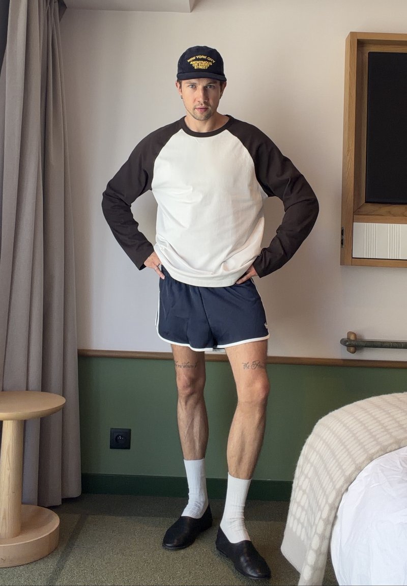 Homme portant un t-shirt à manches longues noir et blanc, un short bleu marine, des chaussettes blanches, des chaussures noires et une casquette noire, debout dans une pièce avec les mains sur les hanches.