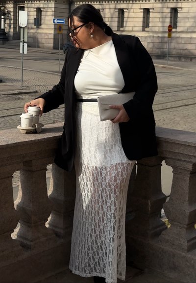 Mujer con falda de encaje blanca, top blanco y blazer negro sujetando un bolso de mano mientras recoge dos tazas de café sobre una barandilla de piedra al aire libre.