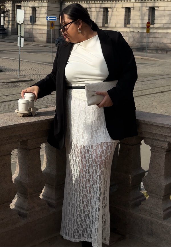 Femme en jupe en dentelle blanche, haut blanc et blazer noir tenant une pochette et ramassant deux tasses de café sur un rebord en pierre extérieur.