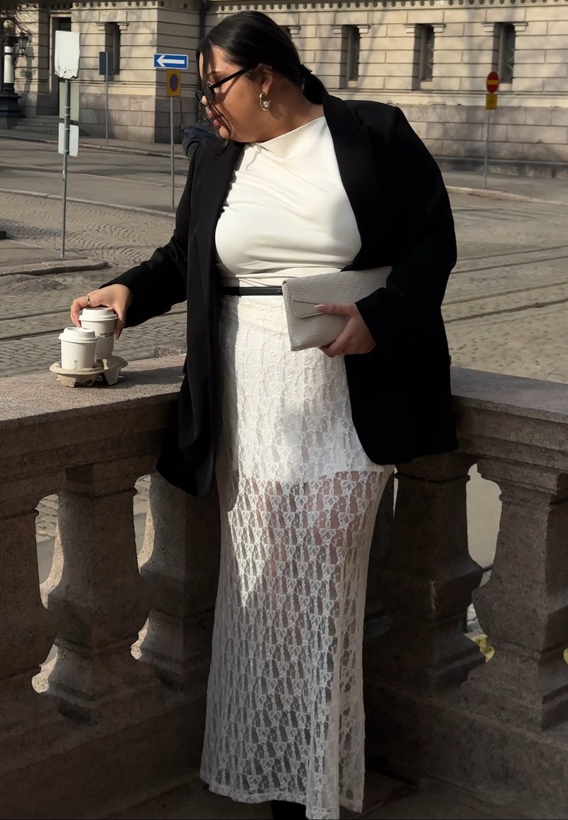 Femme portant une jupe en dentelle blanche, un haut blanc et un blazer noir, tenant une pochette et ramassant deux tasses de café sur une balustrade en pierre en plein air.