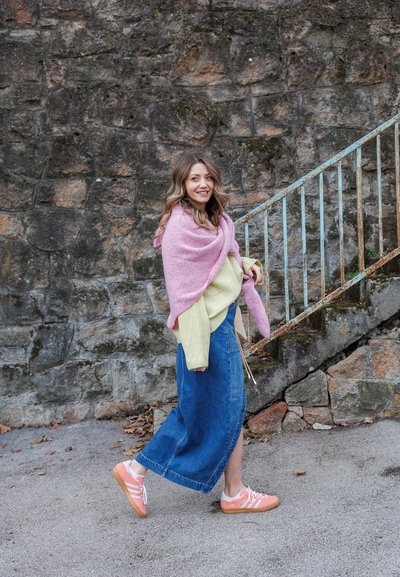 Chal rosa sobre un suéter de punto verde claro, combinado con una falda de mezclilla larga y zapatillas rosas. De pie cerca de una pared de piedra y escaleras de metal.