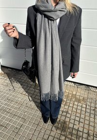 Personne portant un blazer foncé, une grande écharpe grise à franges, un jean foncé, des chaussures noires pointues, tenant un petit sac à main noir avec une bandoulière chaîne.