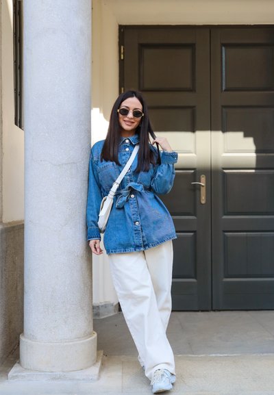 Mujer con chaqueta vaquera y pantalones blancos, de pie y apoyada contra una columna de piedra frente a unas puertas dobles oscuras.