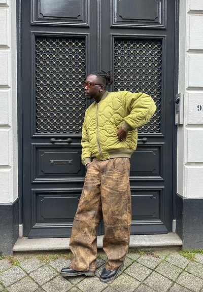 Chaqueta bomber acolchada verde oliva, pantalones marrones sueltos con un acabado texturizado y zapatos negros. Puerta de fondo con un patrón oscuro.