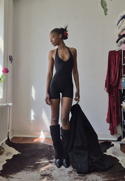 Mujer con un mono negro sin mangas y botas de piel sostiene un abrigo negro, de pie en una habitación con una ventana y ropa colgada.