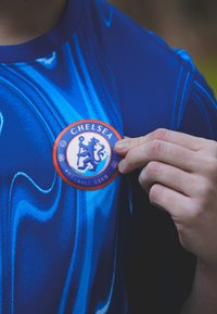 Blue athletic shirt with a wave pattern, featuring a round Chelsea FC badge in orange and blue, held by a hand.