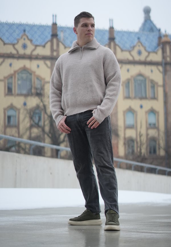 Giovane uomo con maglione beige e jeans scuri in piedi su un marciapiede bagnato con un edificio storico sullo sfondo, guarda di lato.