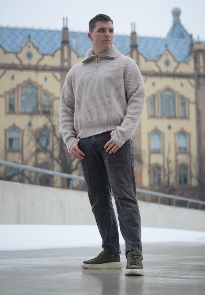 Jeune homme en pull beige et jean foncé debout sur un trottoir mouillé avec un bâtiment historique en arrière-plan, regardant sur le côté.
