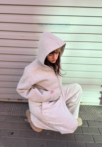 Personne en sweat à capuche beige et pantalon accroupie contre un mur en métal ondulé, regardant vers le bas, les mains dans les poches du sweat à capuche, portant des bottes couleur sable.