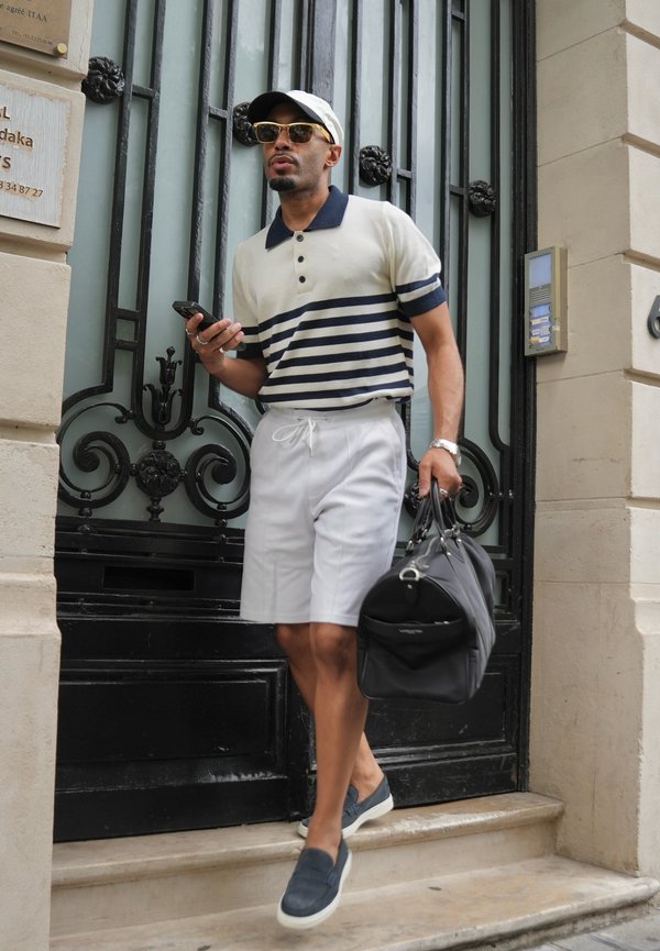 Hombre con gafas de sol, camisa de rayas, pantalones cortos blancos y gorra, bajando las escaleras sosteniendo una bolsa negra y un smartphone junto a una puerta decorativa.