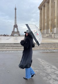 Must kasukas karusnahast detailidega, sinised laia säärega teksad ja must nokamüts, kes hoiab läbipaistvat vihmavarju. Taustal on Eiffel'i torn.