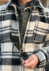 Personne portant une veste à carreaux noire et blanche par-dessus une chemise en velours côtelé verte, tenant des lunettes de soleil à monture noire avec un bandana noir autour du cou.