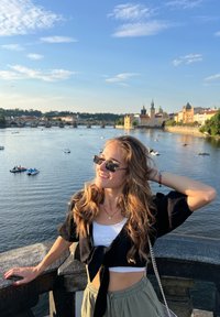 A person wearing a black cropped top and light green pants poses by a river, with sunglasses and jewelry. The water reflects the sky.