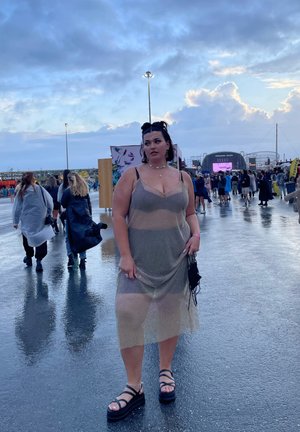 Vestido de malla metálica transparente en dorado con una silueta ajustada, superpuesto sobre ropa interior negra. Sandalias de plataforma negras y un pequeño bolso.