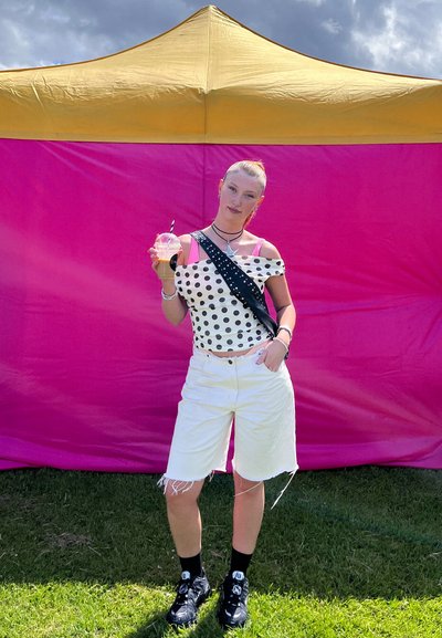 Top blanco con lunares, pantalones cortos blancos, zapatillas negras, sosteniendo una bebida. El fondo presenta una carpa amarilla y rosa. Césped bajo los pies.