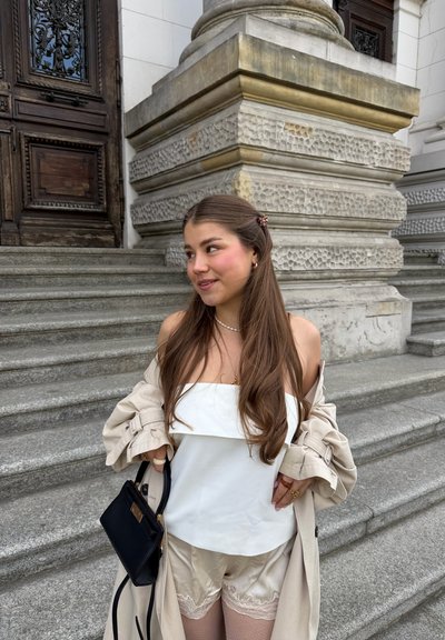 Joven mujer con cabello largo, vestida con una camiseta blanca, abrigo beige y pantalones cortos, de pie en escalones de piedra junto a la entrada ornamentada de un edificio, sosteniendo una bolsa negra.