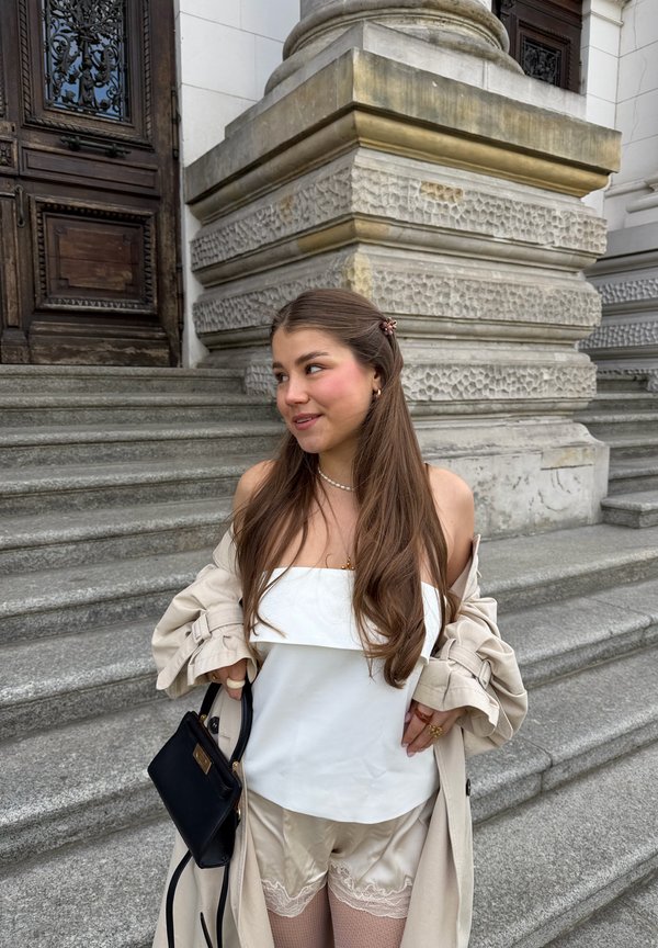 Jeune femme aux longs cheveux, portant un haut blanc, un manteau beige et un short, debout sur des marches en pierre près de l'entrée ornée d'un bâtiment, tenant un sac noir.
