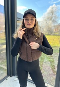 Hnědá péřová vesta s vysokým límcem, zipem vpředu a dvěma bočními kapsami, nošena přes černý dlouhý rukáv a legíny. Na hlavě �černá čepice.