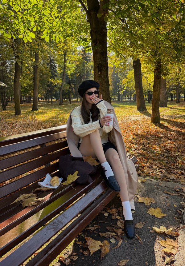 Une femme portant un béret et des lunettes de soleil est assise sur un banc de parc et tient une tasse au milieu des feuilles d’automne.