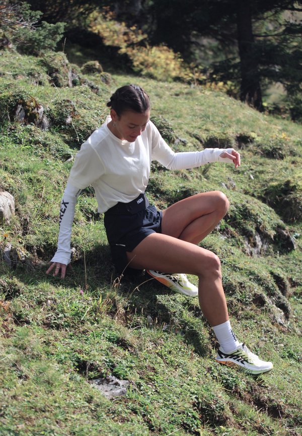 Una mujer vestida para correr está sentada en una ladera cubierta de hierba, apoyándose con una mano y mirando hacia el sendero.