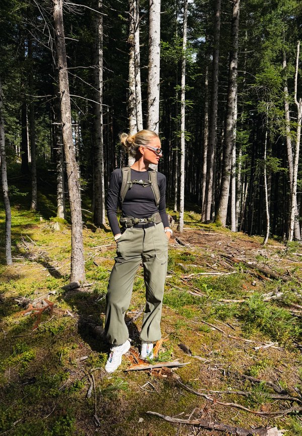 Femme portant des lunettes de soleil et un équipement de randonnée, debout dans une forêt baignée de soleil avec de grands arbres et de la mousse verte au sol.