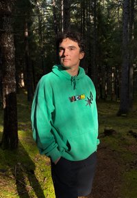 Hanorac turquoise cu șiret și logo, purtat peste pantaloni scurți negri. Textură vizibilă și țesătură netedă, așezat pe un fundal de pădure.