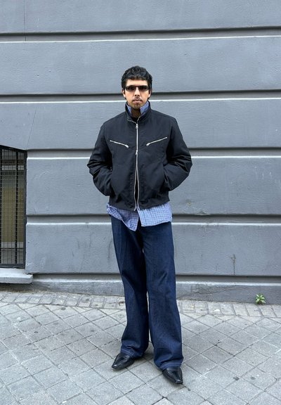 Hombre con chaqueta negra, camisa azul a cuadros, vaqueros anchos oscuros y gafas de sol, apoyado contra una pared gris con las manos en los bolsillos.