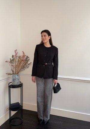 Mujer con blazer negro abotonado con cinturón, pantalones grises, sosteniendo un pequeño bolso negro, de pie junto a un soporte para plantas negro con flores secas.