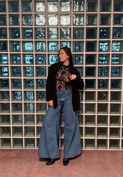 Cárdigan negro de manga larga sobre un body negro estampado, pantalones vaqueros de pierna ancha en azul y botas negras hasta el tobillo. Pared de bloques de cristal en el fondo.