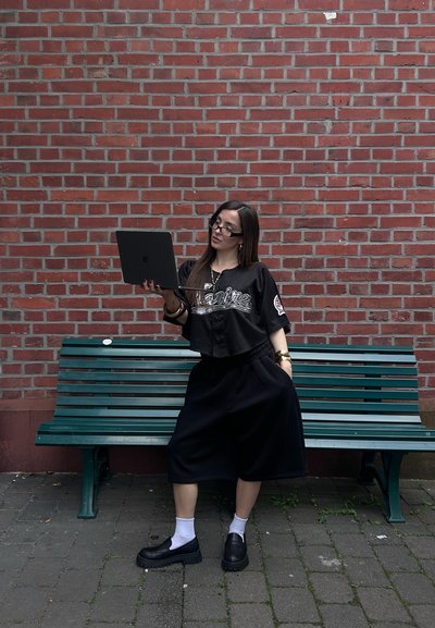 Jersey negro corto con letras plateadas, shorts negros holgados, zapatos negros, calcetines blancos, sosteniendo un portátil, sentado en un banco verde. Fondo de pared de ladrillo.