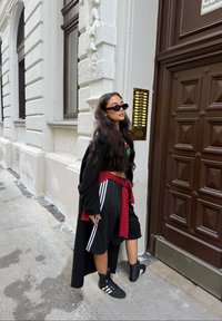 Svart lång kappa över en svart croppad grafisk t-shirt, röd och svart rutigt slips vid midjan, svarta shorts med vita ränder, svarta sneakers.