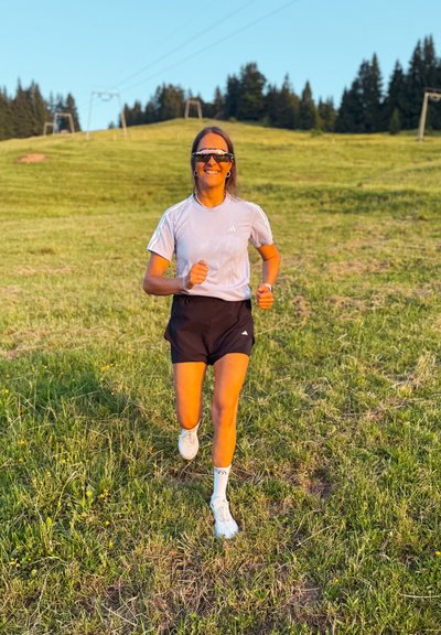 Conjunto de ropa atlética para correr que incluye una camiseta de manga corta gris claro, pantalones cortos negros, zapatillas de correr blancas y calcetines largos blancos, sobre un terreno cubierto de hierba.