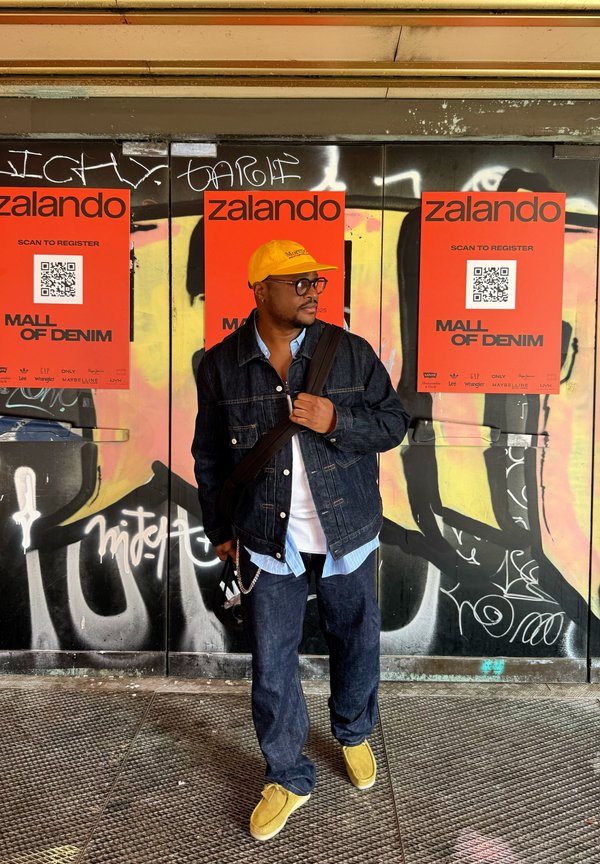 Denim jacket over a light blue shirt, dark jeans, and yellow slip-on shoes. Black glasses and a yellow cap. The background features graffiti and posters.