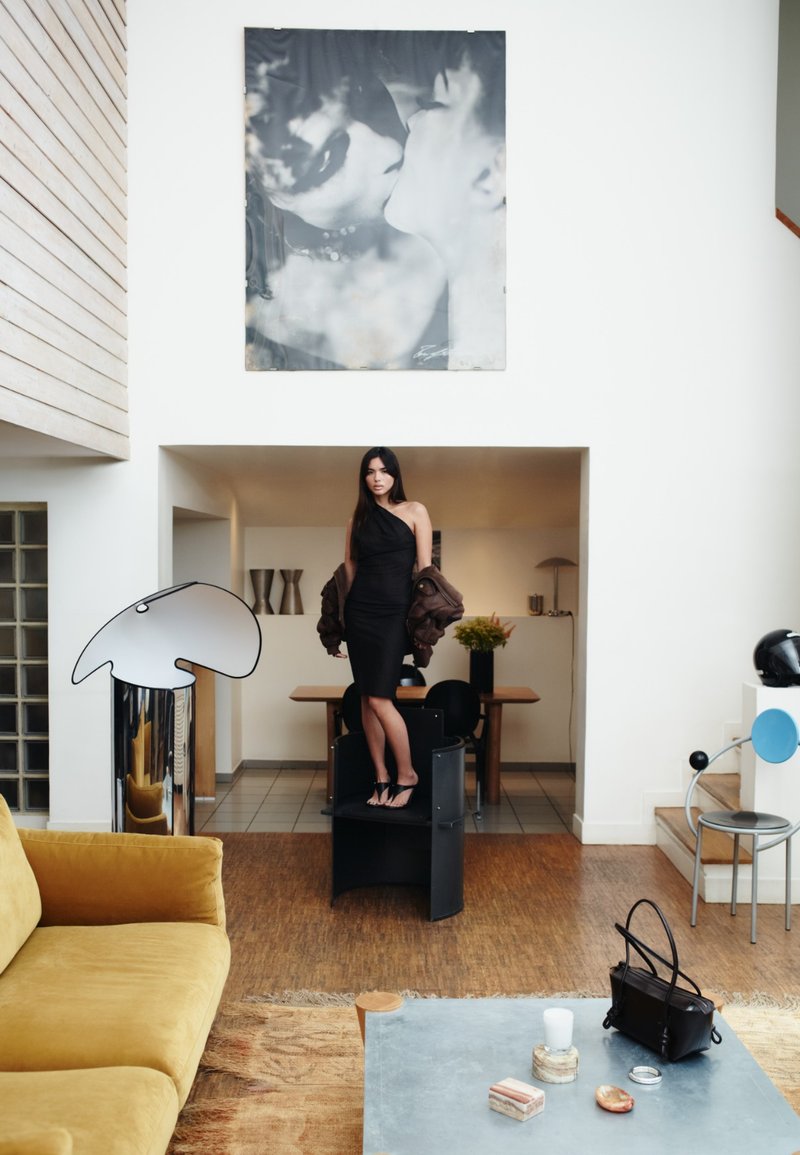 Vestido preto, casaco oversized castanho, de pé num pedestal cilíndrico preto. A decoração moderna inclui um sofá amarelo e arte abstracta. Mesa de vidro com acessórios.