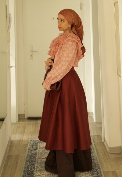 Mujer con blusa rosa con volantes y estampado floral, hijab marrón y falda larga burdeos, de pie de lado en un pasillo con una alfombra en el suelo.