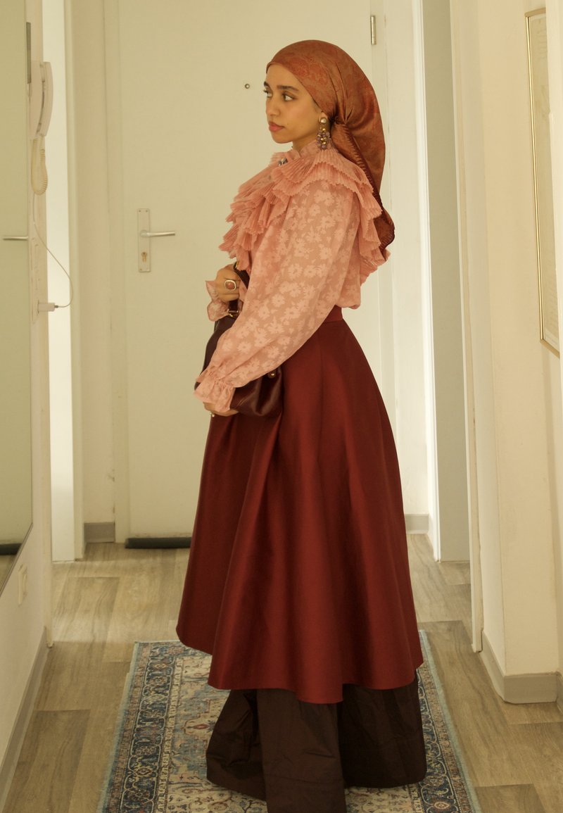Femme en blouse rose à fleurs avec volants, hijab marron et longue jupe bordeaux, debout de profil dans un couloir avec un tapis au sol.