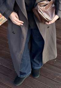 Personne portant un trench-coat sombre, un jean à jambes larges et des chaussures noires, tenant un sac en cuir brun texturé tout en se tenant sur un plancher en bois.