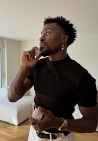 Homme appliquant de la crème sur son visage à l'intérieur, tenant un pot, vêtu d'une chemise noire, d'un pantalon blanc, portant une montre en or et une boucle d'oreille.