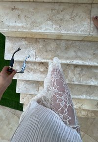 Μαύρα γυαλιά ηλίου με καπιτονέ σκελετό κρατημένα πάνω από ανοιχτόχρωμες μαρμάρινες σκάλες, συνοδευόμενα από ένα άσπρο κομμάτι δαντέλας με λου�λουδάτα σχέδια.