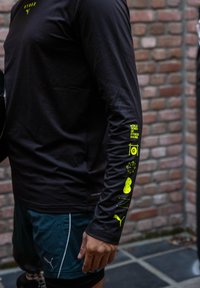 Must pika varrukaga särk, mille vasakul varrukal on neoonkollased graafikud ning mille lõige on kitsas. Kandmisega koos on türkiissinised lühikesed püksid ja sportlik välimus.