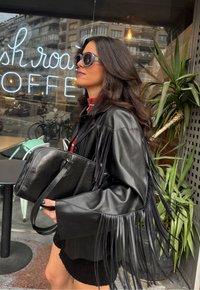 Μαύρο δερμάτινο μπουφάν με φούντες, σε συνδυασμό με μια μαύρη τσάντα. Το μοντέλο έχει μακριά, κυματιστά μαλλιά και φοράει oversized γυαλιά ηλίου. Καθαρό αστικό φόντο.