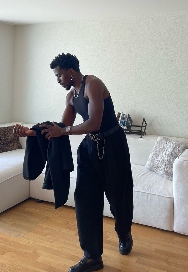 Mężczyzna w czarnym tank topie i szerokich spodniach trzyma czarną kurtkę, stoi w salonie z białą sofą i drewnianą podłogą.