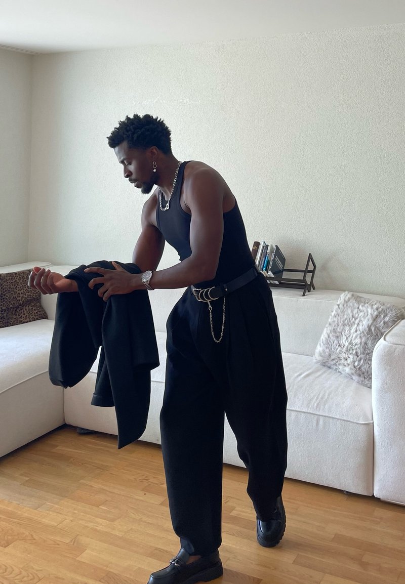 Mies mustassa toppatopissa ja leveälahkeisissa housuissa, jolla on musta takki kädessään, seisoo olohuoneessa, jossa on valkoinen sohva ja puulattia.