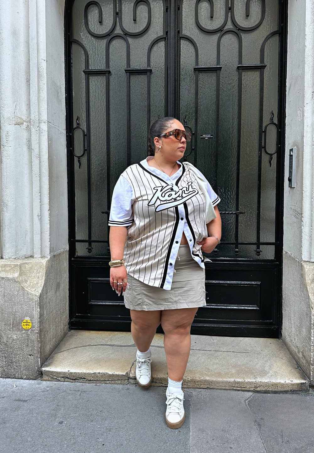 Overmaatse gestreepte honkbaltrui in wit en zwart, gecombineerd met een beige minirok en witte sneakers. Zonnebril en gestapelde armbanden.