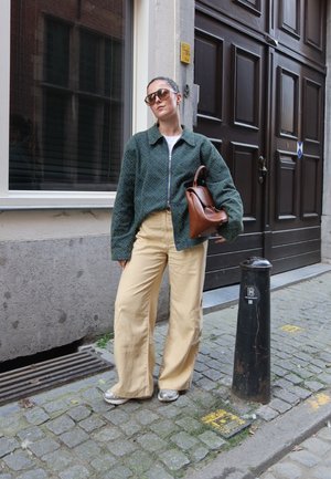 Chaqueta verde texturizada con cremallera, camiseta blanca, pantalones anchos beige, zapatillas plateadas y bolso de cuero marrón. Entorno urbano con adoquines.